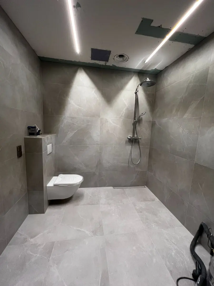 Færdigt badeværelse med store gråmarmorerede fliser på gulv og vægge, linjeafløb i gulvet, flisebeklædt indbygning ved det væghængte toilet og lysstriber i det nedsænkede loft