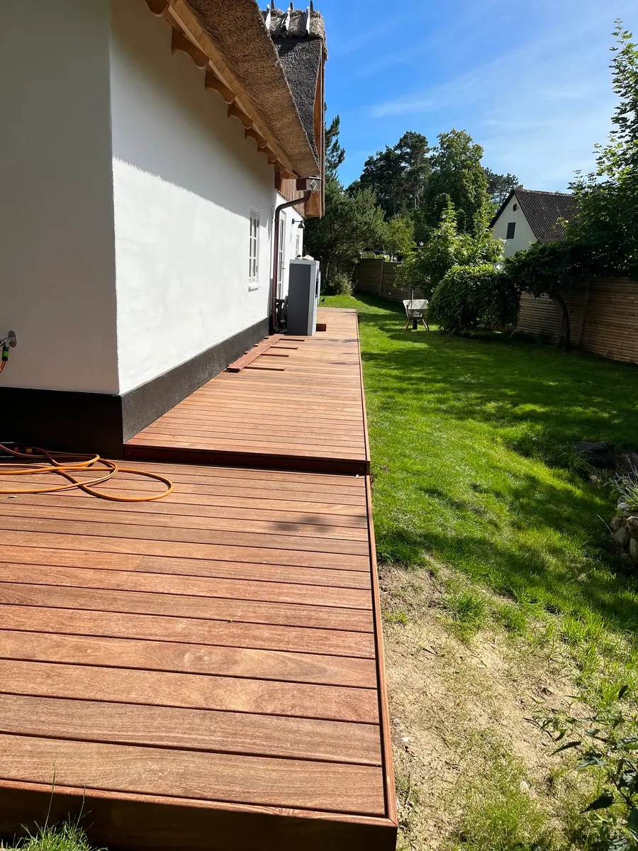Terrasse i hårdttræ ført langs hele den hvide facade på et stråtækt hus, med græsplæne og hæk ved siden af